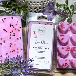 May include: A set of pink heart-shaped wax melts in a clear plastic container. The wax melts are labeled "Love Potion Wax Melt Bar" with the scents "Rose, Grapefruit, Chamomile". The label also states "100% Natural Soy Wax, Hand Poured in Small Batches, 3 oz, Not for Consumption".