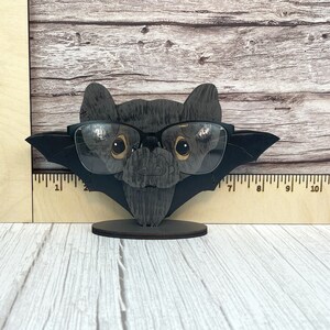 May include: A black wooden bat with a pair of black framed glasses perched on its nose. The bat is standing on a small wooden base.