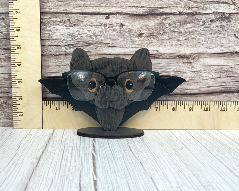 May include: A black wooden bat with a pair of black framed glasses perched on its nose. The bat is standing on a small wooden base.