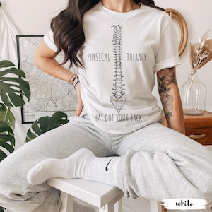 May include: A white short-sleeved t-shirt featuring a black line drawing of a human spine centered on the chest. The text "PHYSICAL THERAPY" is printed above the spine, and "HAS GOT YOUR BACK" is printed below the spine in black lettering.