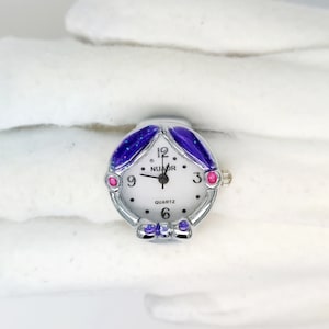 May include: A silver ring watch with a white face and black hands. The watch has a purple and pink design with glitter accents. The watch face says "NUMOR QUARTZ".