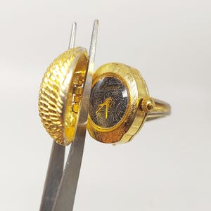 Könnte beinhalten: Eine goldfarbene Armbanduhr mit schwarzem Zifferblatt und einem dekorativen Muster. Die Uhr hat die Form eines Rings und einen Klappdeckel.