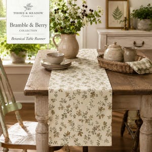 May include: A cream-colored table runner featuring a delicate brown and green botanical vine pattern. The runner is laid across a wooden table, topped with a ceramic pitcher filled with flowers, a small bowl, and a woven basket with a pitcher.