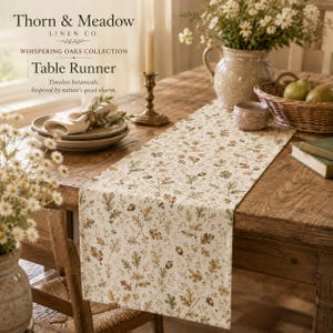 May include: A rectangular table runner featuring a repeating pattern of brown and green oak leaves and acorns on a cream-colored fabric. The runner is laid flat on a wooden table, showcasing a botanical design with natural earth tones.