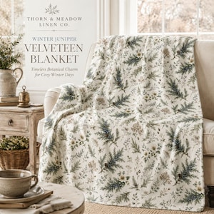 May include: A cream-colored velveteen blanket featuring a botanical print of green juniper branches, pine needles, and small white flowers. The text on the image reads: THORN & MEADOW LINEN CO. WINTER JUNIPER VELVETEEN BLANKET Timeless Botanical Charm for Cozy Winter Days.