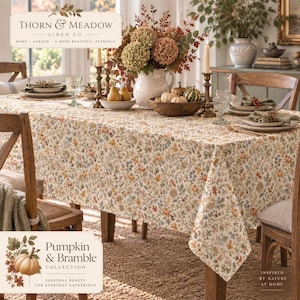 May include: A rectangular tablecloth featuring a dense, repeating pattern of small orange pumpkins, autumn leaves, and wildflowers in muted earth tones. The fabric drapes over a dining table set with a ceramic pitcher, bowls of fruit, and place settings.