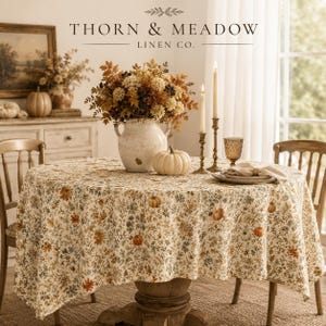 May include: A round tablecloth featuring a cream background with a dense botanical print of orange, brown, and green flowers, leaves, and small pumpkins. The table is set with a white ceramic vase of dried flowers and a white pumpkin.