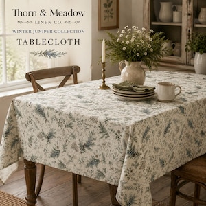 May include: A rectangular tablecloth featuring a botanical print of green juniper branches and small flowers on a cream background. The fabric drapes over a wooden table, which is set with a ceramic vase of white flowers, a candle, and a mug.