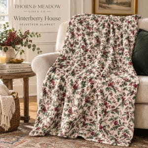 May include: A cream-coloured velveteen blanket featuring a repeating pattern of dark green holly leaves and clusters of red winterberries. The text on the image reads Thorn & Meadow Linen Co. Winterberry House Velveteen Blanket.