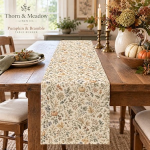 May include: A rectangular table runner featuring a cream-coloured fabric with an intricate botanical print of small pumpkins, autumn leaves, and bramble vines in muted shades of orange, brown, green, and blue, labelled Thorn & Meadow Pumpkin & Bramble.