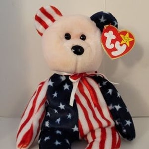 May include: A plush teddy bear wearing a red, white, and blue American flag patterned outfit. The bear has a pink nose and black eyes. The bear is sitting on a white surface. The bear has a Ty tag attached to its ear.