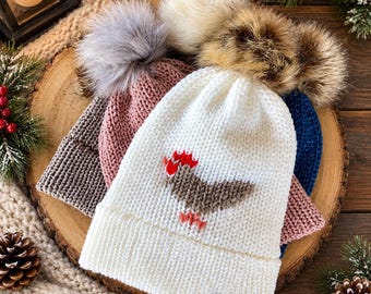 May include: A collection of four knit beanies with faux fur pom-poms in white, pink, grey, and blue. The white beanie features a brown and red chicken graphic on the front and a small brown leather-like tag at the bottom edge.