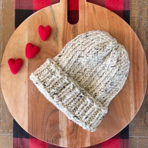 May include: A chunky knit beanie in cream with black flecks, folded at the brim. Three small red felt hearts are arranged near the hat on a wooden cutting board. The board rests on a red and black plaid surface.