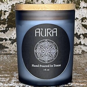 May include: A black and white candle jar with a wooden lid. The jar has a label that says "AURA" and "Hand Poured in Texas" with a decorative design. The candle is 7 fl. oz.