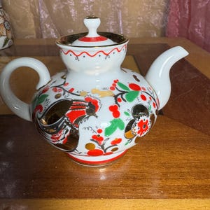 Könnte beinhalten: Weiße Porzellan-Teekanne mit rotem, grünem und goldenem Blumendesign. Die Teekanne hat einen runden Korpus und einen Deckel mit einem Knopf.