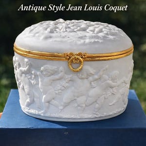 May include: White Limoges cherub jewelry box with gold trim. The box features a relief design of cherubs and is labeled "CHERUB JEWELRY BOX" and "Antique Style Jean Louis Coquet". The box is made of biscuit porcelain.