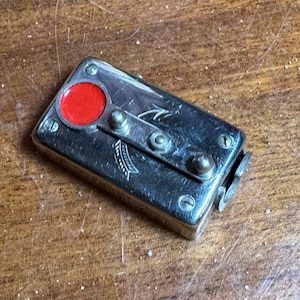 Può includere: Un dispositivo in metallo argentato con un cerchio rosso e due piccoli pomelli in metallo. Il dispositivo ha un design decorativo sulla parte superiore.