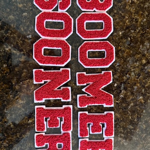 May include: Red and white embroidered patches spelling "SOONER BOOMER". The letters are in a varsity style with a white border. The patches are likely for a jacket or bag.