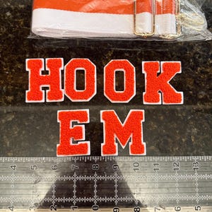 May include: An orange and white striped bag strap with gold-colored hardware is displayed above embroidered patches spelling "HOOK EM". The patches are orange with white borders. A ruler is in the foreground.