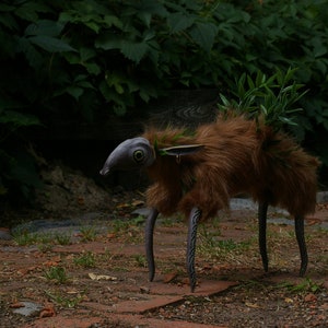 May include: A whimsical creature made of fur and metal with a fish-like head and green foliage growing on its back. The creature has long, thin legs and is standing on a path of brown bricks.