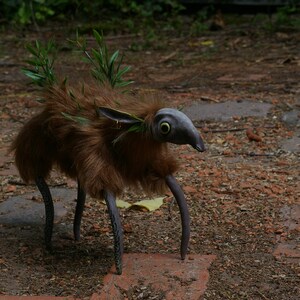 May include: A brown, furry creature with long, spindly legs and a long, pointed snout. The creature has large, green eyes and is adorned with green leaves. It is standing on a dirt path.