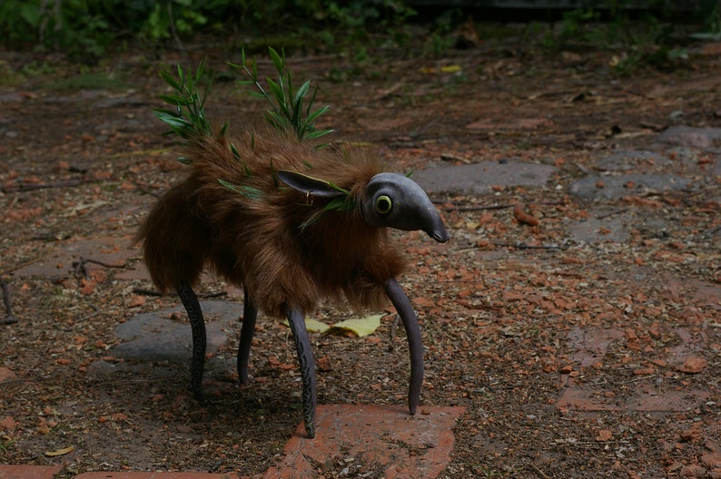 May include: A brown, furry creature with long, spindly legs and a long, pointed snout. The creature has large, green eyes and is adorned with green leaves. It is standing on a dirt path.