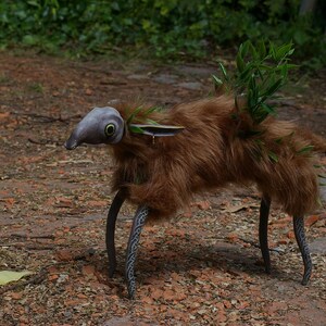 May include: A whimsical creature made of brown fur, with a gray, bird-like head and large green eyes. It has long, black, metal legs with intricate designs. The creature is standing on a path covered in brown leaves and rocks.