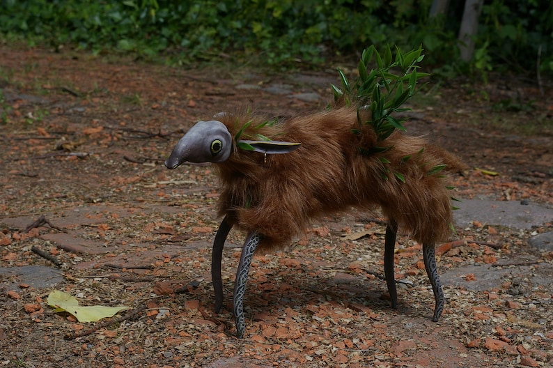 May include: A whimsical creature made of brown fur, with a gray, bird-like head and large green eyes. It has long, black, metal legs with intricate designs. The creature is standing on a path covered in brown leaves and rocks.