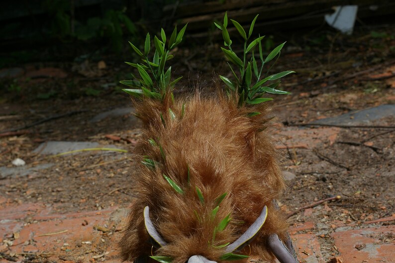 May include: A brown furry creature with green leafy branches on top of its head. The creature is standing on a dirt and brick surface.