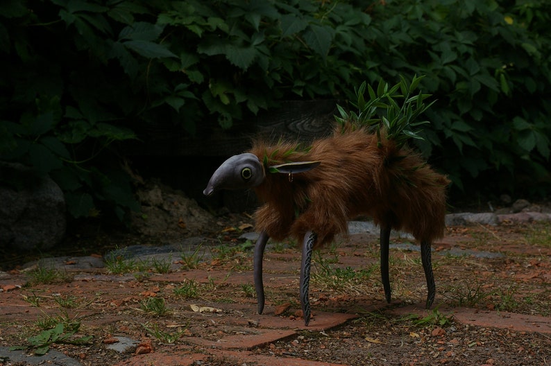 May include: A whimsical creature made of fur and metal with a fish-like head and green foliage growing on its back. The creature has long, thin legs and is standing on a path of brown bricks.