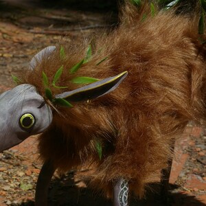 May include: A whimsical, furry creature with a long, gray snout and large, yellow eyes. It has brown fur and green leaves woven into its fur. The creature has a long, thin tail and stands on four legs.