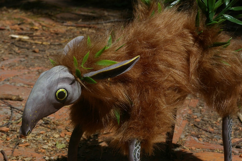 May include: A whimsical, furry creature with a long, gray snout and large, yellow eyes. It has brown fur and green leaves woven into its fur. The creature has a long, thin tail and stands on four legs.
