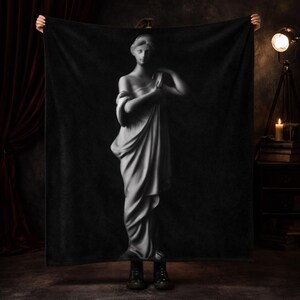 May include: A rectangular black throw blanket featuring a large, centered, high-contrast grayscale print of a classical marble statue of a figure draped in flowing fabric. The design highlights the detailed folds of the garment and the statue's form.