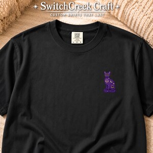 May include: A black short-sleeved t-shirt featuring a small purple cat graphic on the chest. The cat design includes celestial symbols like stars, moons, and an eye. The shirt has a Comfort Colors label at the neck and the text SwitchCreek Craft.
