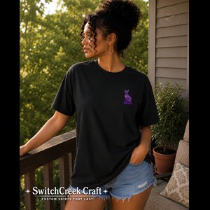 May include: A black short-sleeve t-shirt featuring a small, purple, celestial-themed cat graphic on the left chest. The cat design includes moon and star patterns in shades of purple and white. The shirt has a crew neckline and a relaxed fit.