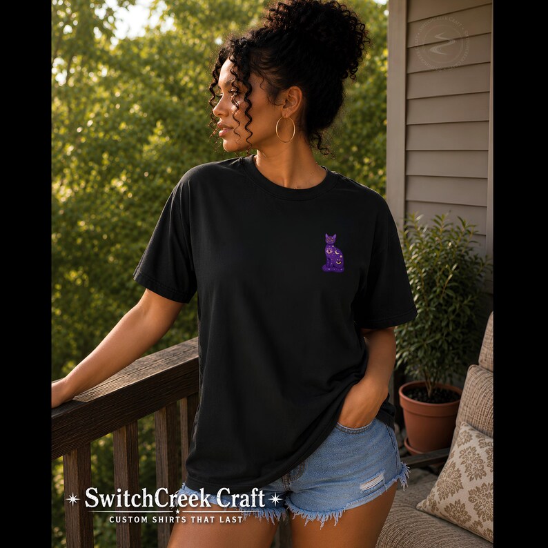 May include: A black short-sleeve t-shirt featuring a small, purple, celestial-themed cat graphic on the left chest. The cat design includes moon and star patterns in shades of purple and white. The shirt has a crew neckline and a relaxed fit.
