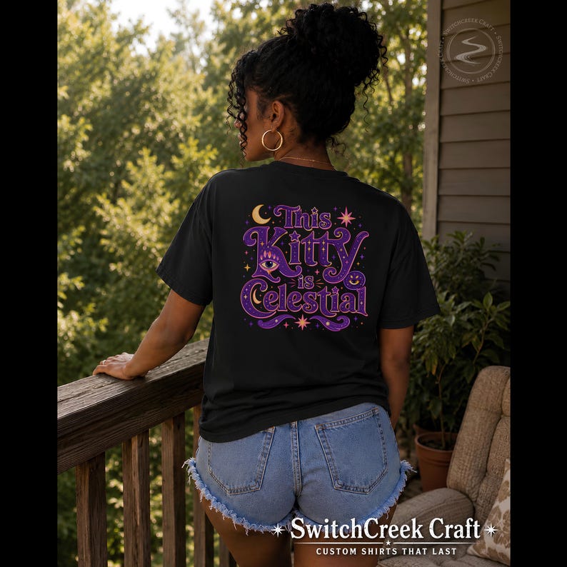 May include: A black t-shirt featuring a graphic on the back with the text This Kitty is Celestial in purple and orange lettering. The design includes a crescent moon, stars, a stylized eye, and a small smiling face, all set against a dark background.