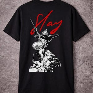 Op de afbeelding: Zwart t-shirt met een witte afbeelding van een krijger met schild en speer, bovenop een wezen. Het woord "Slay" staat in rode letters erboven. Het shirt hangt aan een houten hanger.