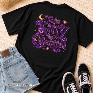 May include: A black short-sleeved t-shirt featuring a graphic print on the back with the text "This Kitty is celestial" in purple and orange. The design includes a stylized eye, crescent moons, and stars in shades of purple, orange, and yellow.
