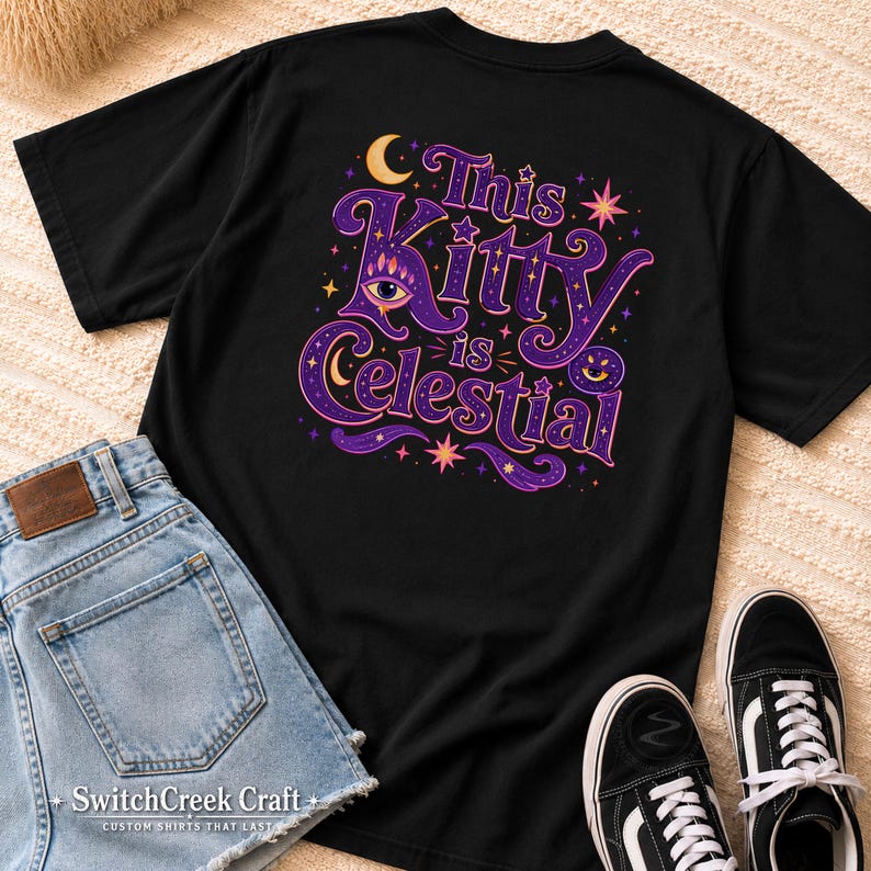 May include: A black short-sleeved t-shirt featuring a graphic print on the back with the text "This Kitty is celestial" in purple and orange. The design includes a stylized eye, crescent moons, and stars in shades of purple, orange, and yellow.