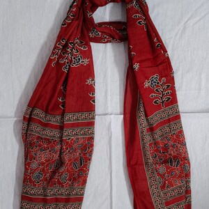 May include: A red scarf with a floral pattern in black and white. The scarf has a border with a repeating design. The scarf is made of a lightweight fabric and is draped on a white surface.