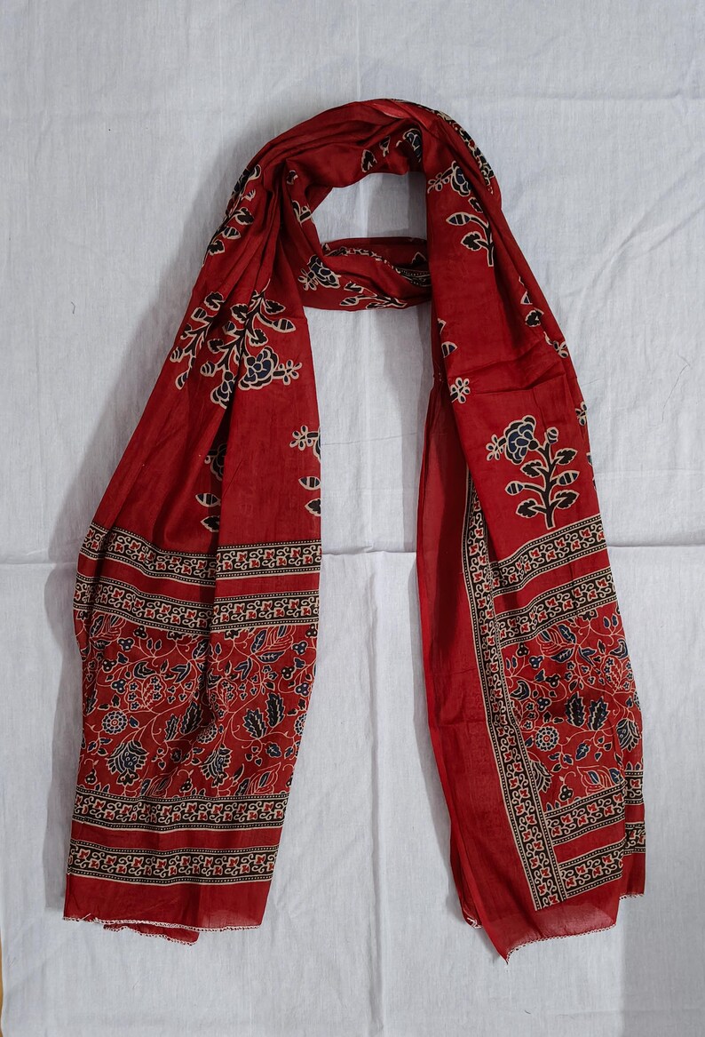 May include: A red scarf with a floral pattern in black and white. The scarf has a border with a repeating design. The scarf is made of a lightweight fabric and is draped on a white surface.