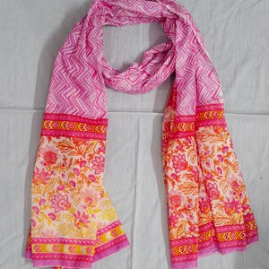 May include: A pink scarf with a white and pink chevron pattern and floral print. The scarf has a border with a repeating design in pink, yellow, and orange. The scarf is made of a lightweight fabric.