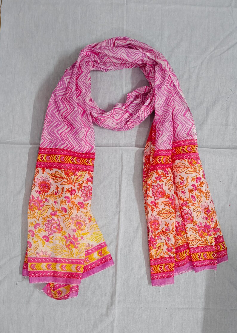 May include: A pink scarf with a white and pink chevron pattern and floral print. The scarf has a border with a repeating design in pink, yellow, and orange. The scarf is made of a lightweight fabric.