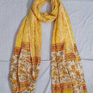 May include: A yellow scarf with a floral and geometric pattern. The scarf features a repeating zigzag design in yellow and orange, with floral borders in white, yellow, and orange. The scarf is made of a lightweight fabric.