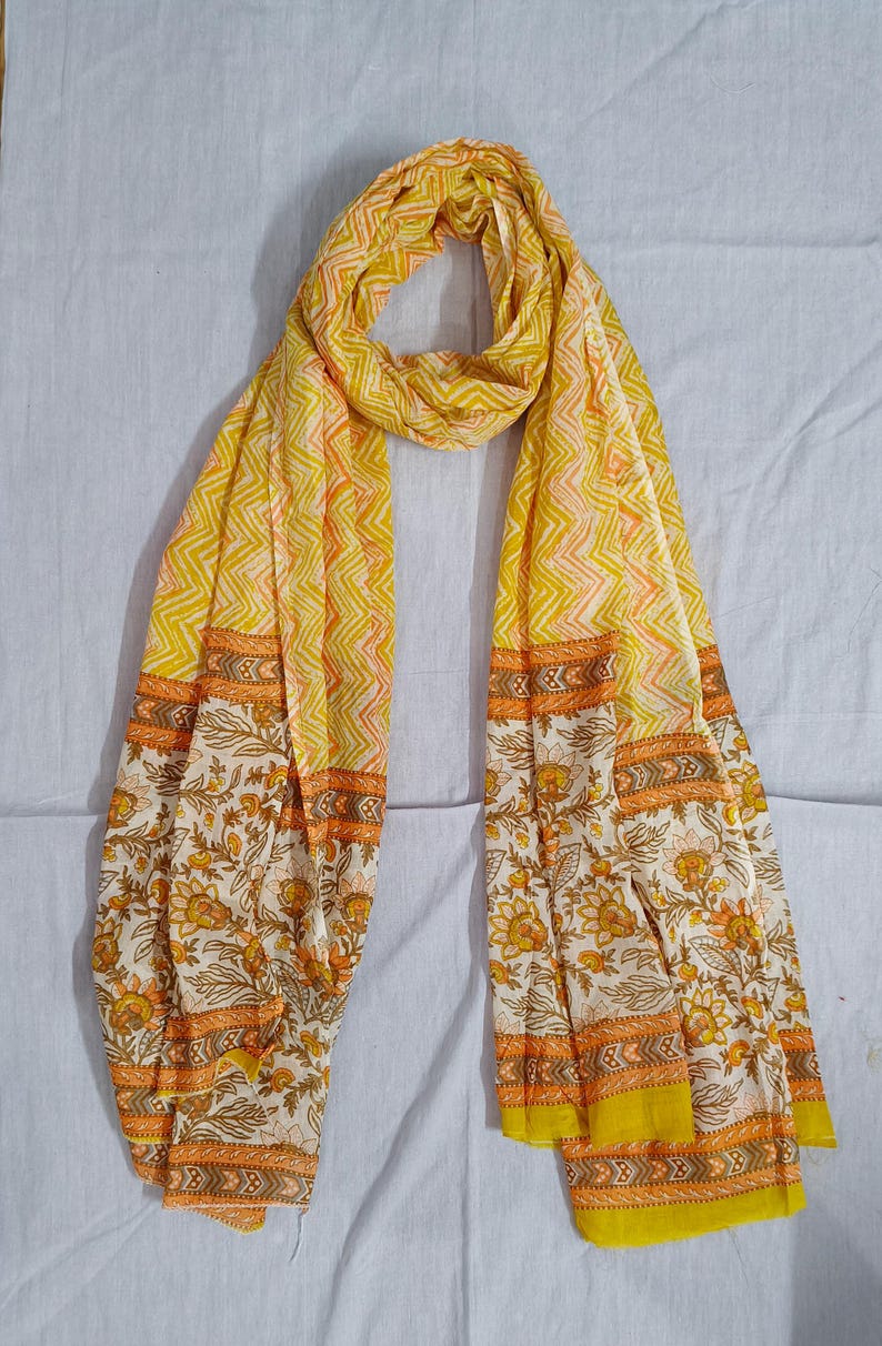 May include: A yellow scarf with a floral and geometric pattern. The scarf features a repeating zigzag design in yellow and orange, with floral borders in white, yellow, and orange. The scarf is made of a lightweight fabric.