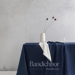 May include: A deep navy blue tablecloth draped over a surface, featuring a white napkin folded on top. A ribbed white vase holds three purple allium flowers. The text Bandichhor Handloom is printed in white in the center of the navy fabric.