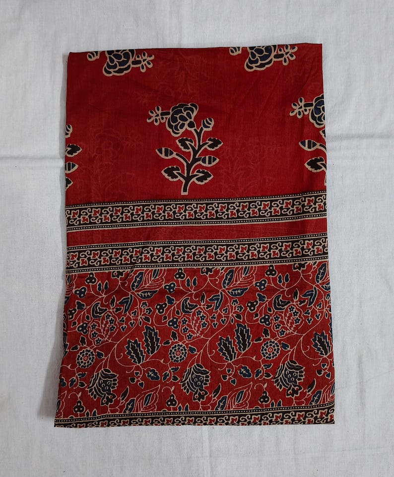 May include: Red fabric with a floral pattern. The fabric features a repeating design of black and white flowers and leaves. A decorative border with a geometric pattern separates the floral design from the solid red background.