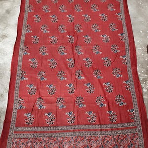 May include: A red textile with a floral pattern. The fabric features repeating blue and white flowers on stems, framed by detailed borders. The borders have intricate designs in red, black, and white.