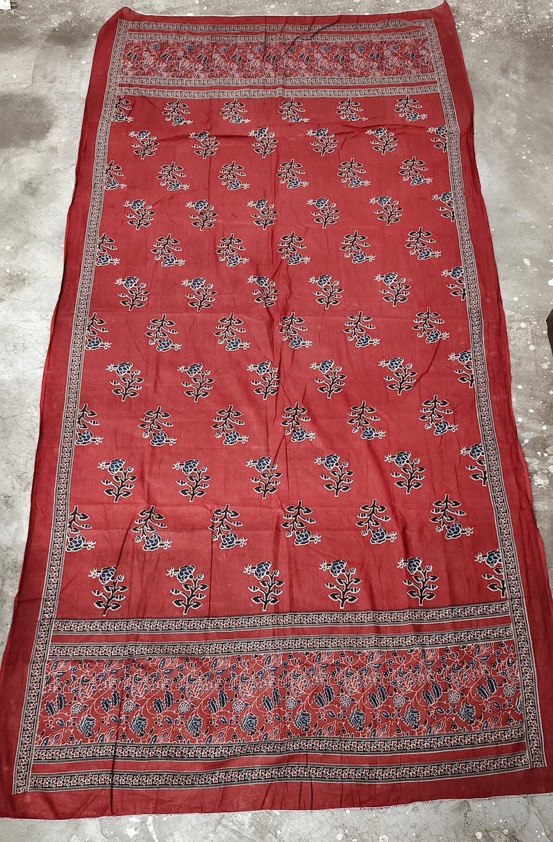 May include: A red textile with a floral pattern. The fabric features repeating blue and white flowers on stems, framed by detailed borders. The borders have intricate designs in red, black, and white.